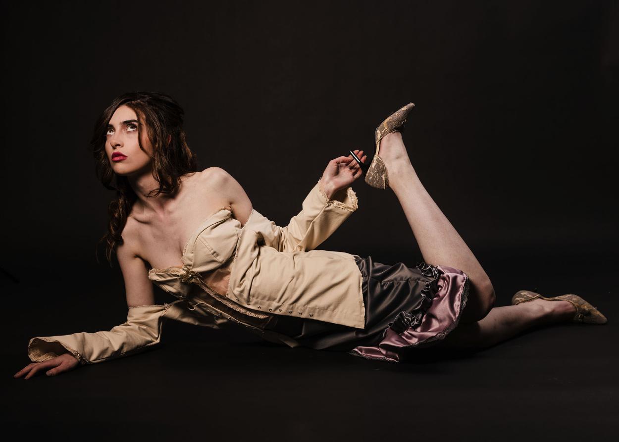 First model lying on the floor, wearing a grey satin skirt and a beige corset with silk and lace details, against a black backdrop
