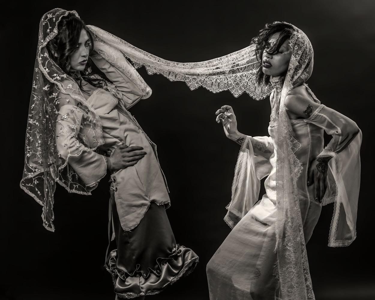 First model wearing a grey satin skirt and a beige corset is pulling the veil of the third model, who is wearing a white bridal gown, against a black backdrop  