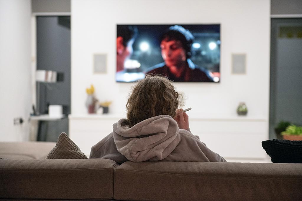 A person in a hoodie sits on a sofa smoking while watching television in a dimly lit living room.