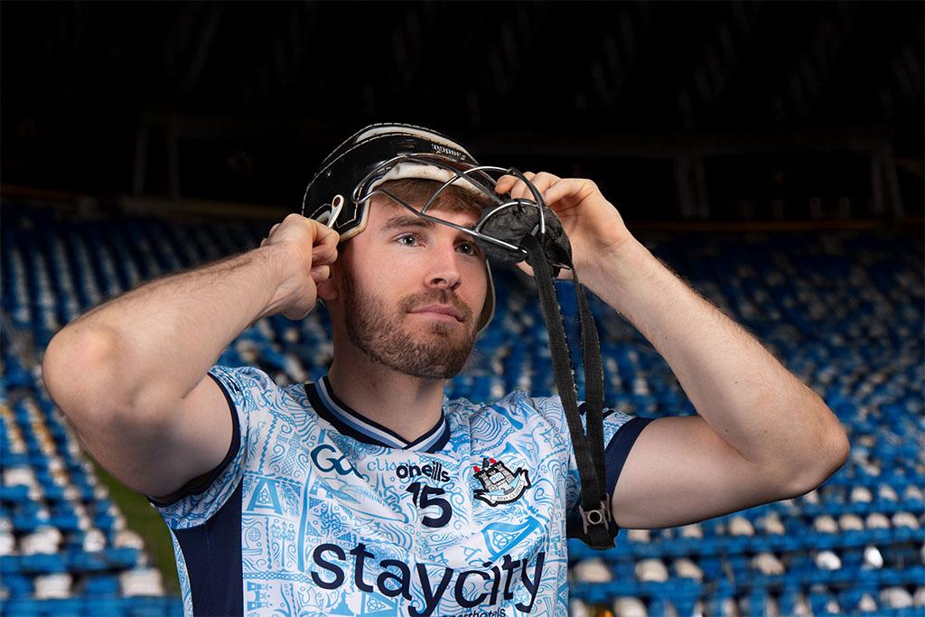A hurler in a Dublin jersey adjusts a helmet while standing in a stadium with rows of blue seats in the background.