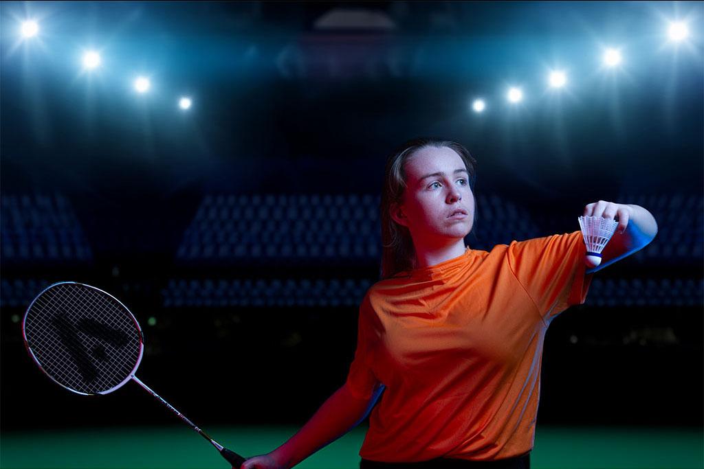 A badminton player in an orange shirt prepares to serve a shuttlecock under dramatic blue and red lighting.