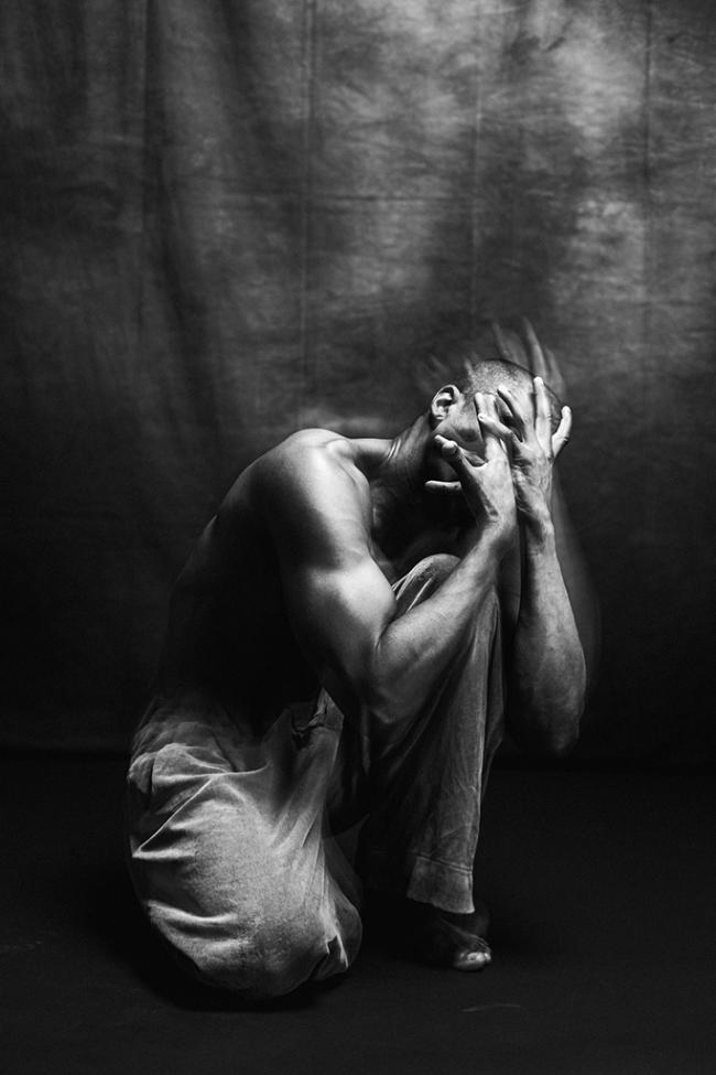 Black and white image of crouched performer on dark stage with head bowed and hands over face.