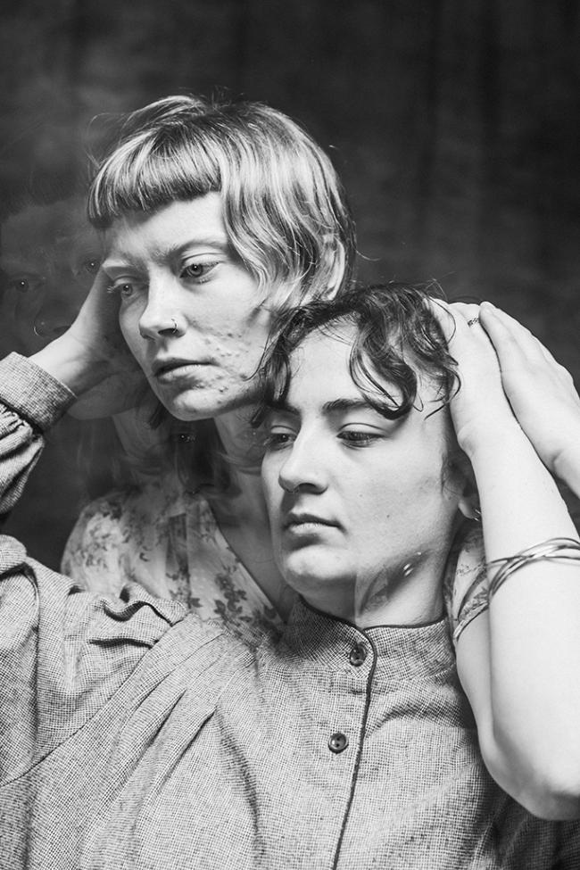 Black and white image of two performers close together, one behind the other, with overlapping double‑exposed faces.