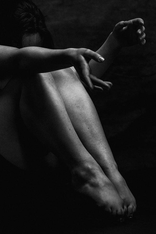 Black and white image of a seated figure with arms extended and legs drawn in, partially obscuring the face.