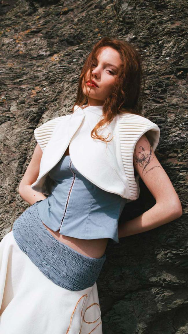 ¾ view model wearing báinín and grey cotton outfit, featuring shirring and tucked fabric manipulation, with rough coastal rock in background.