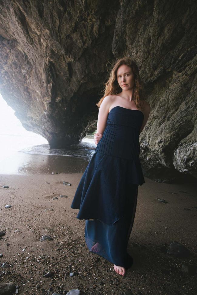 Model wearing denim corset and denim asymmetric skirt over a chiffon underskirt with rough coastal rock in background.