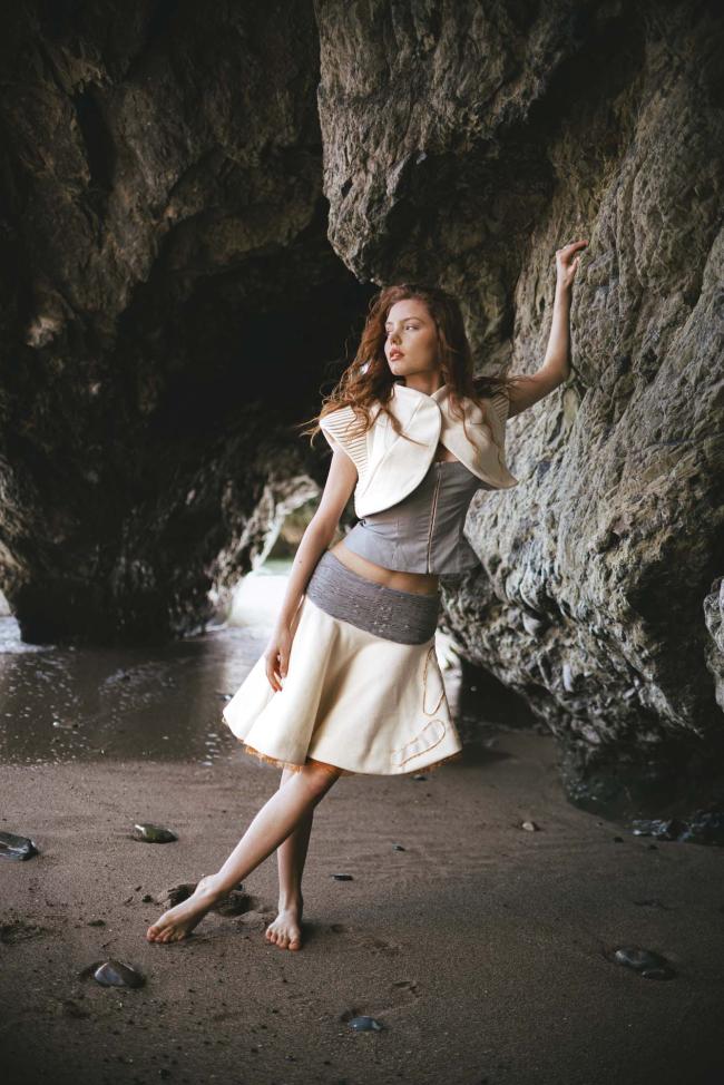  Full-length model wearing báinín and grey cotton outfit, featuring shirring and tucked fabric manipulation, with rough coastal rock in background.