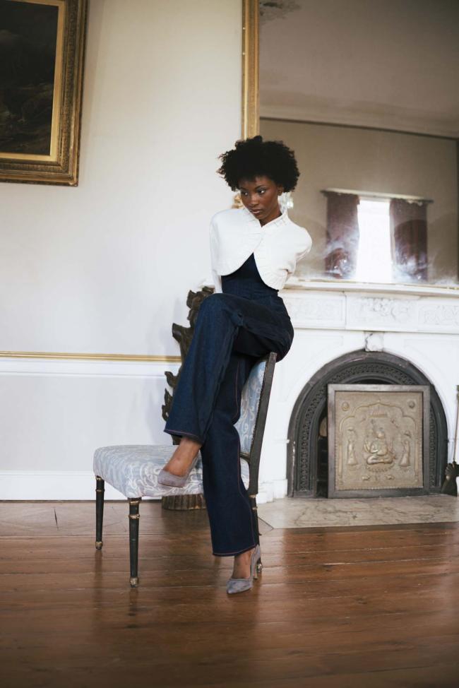 Model wearing denim trousers, corset and báinín jacket, posed sitting on the back of a chair.
