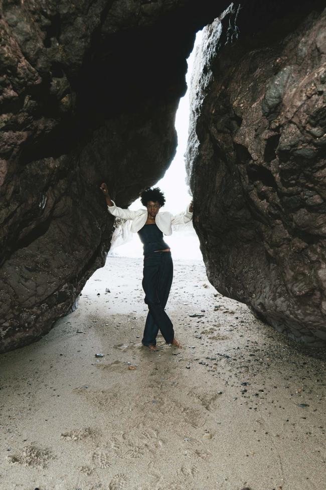 Model wearing denim trousers, corset and báinín jacket, with rough coastal rock in background.
