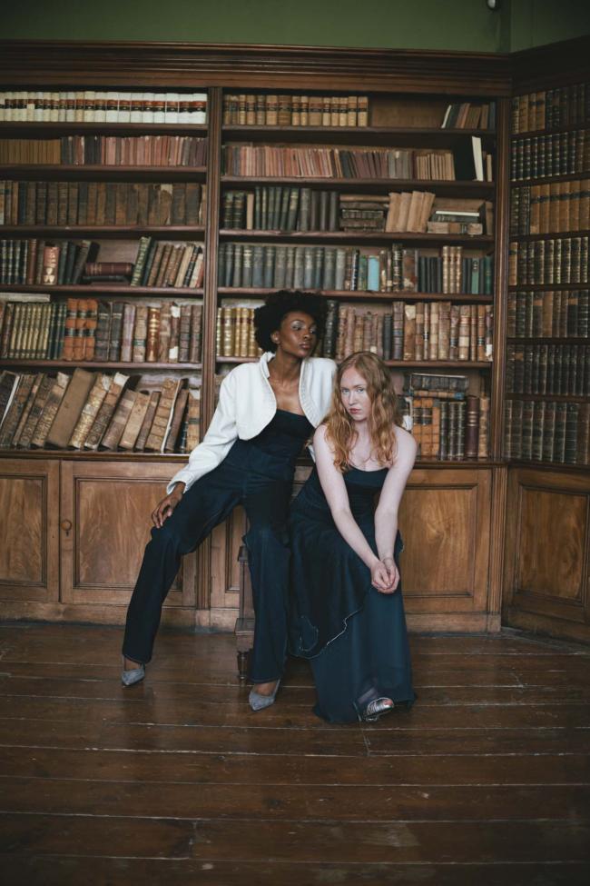 Two models wearing denim trousers, corset and báinín jacket, and denim corset and denim asymmetric skirt over a chiffon underskirt, posed in a library.