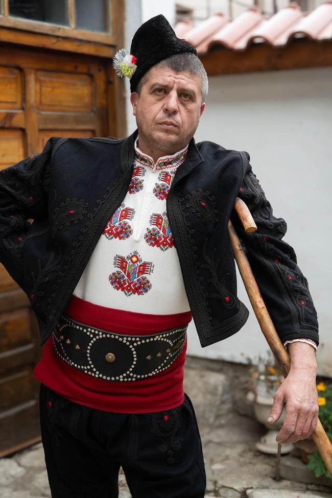  A man in traditional embroidered clothing and a black hat stands holding a wooden staff in a courtyard.