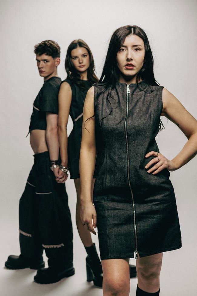 Two female models wear black denim dresses, and a male model wears a black denim top with black denim jeans.