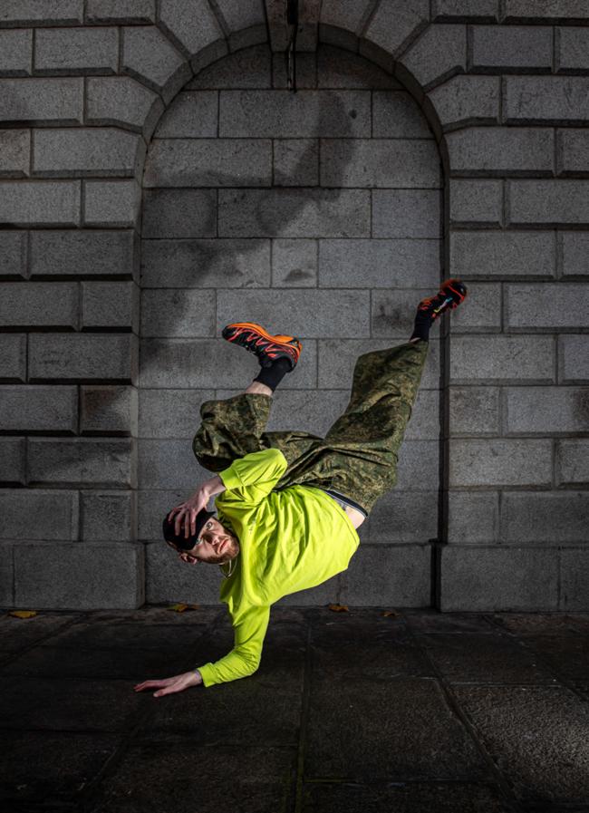A person in a neon yellow hoodie performs a one-handed freeze, legs kicked up in the air, against a gray stone archway. 
