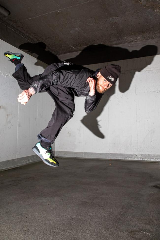 A person in all-black clothing mid-jump, body horizontal with legs kicked up, casting a dramatic shadow on a concrete wall.