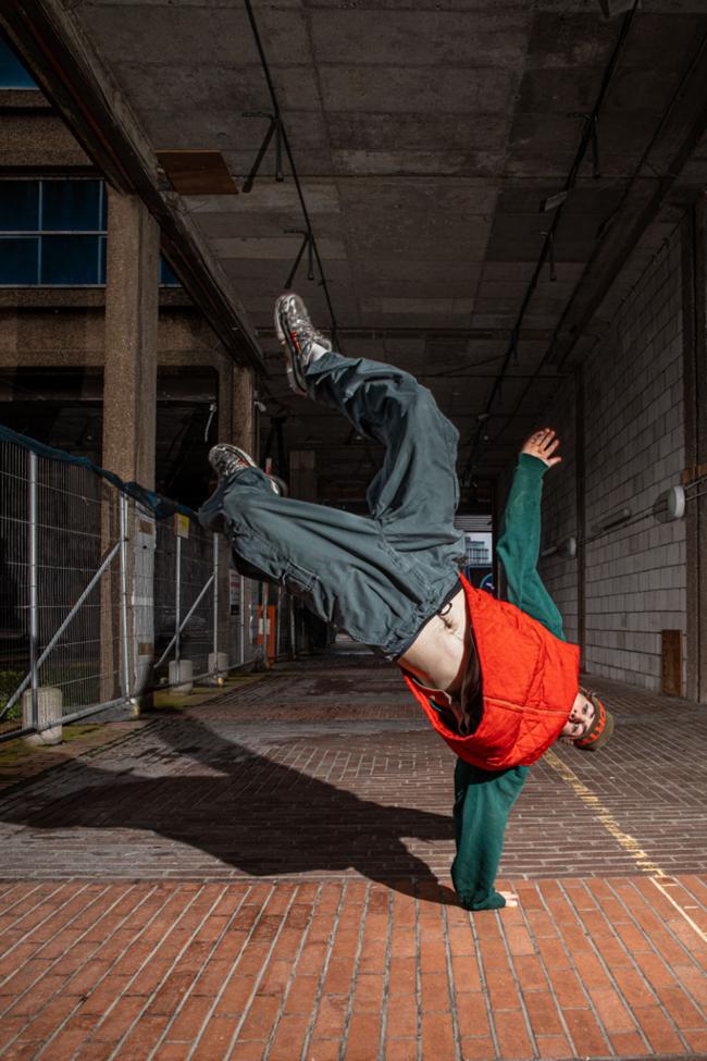 A person in an orange puffer vest and green hoodie performs a one-handed freeze in an urban walkway, legs kicked high toward the ceiling.