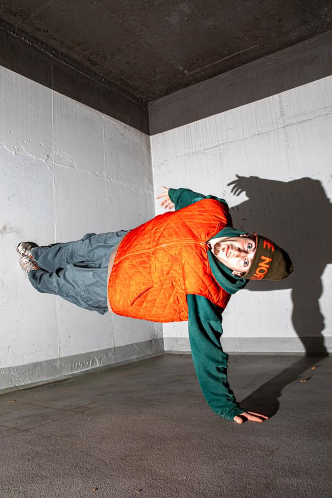 A person in an orange vest and green outfit balances on one hand, body angled sideways, casting a sharp shadow on a white concrete wall.