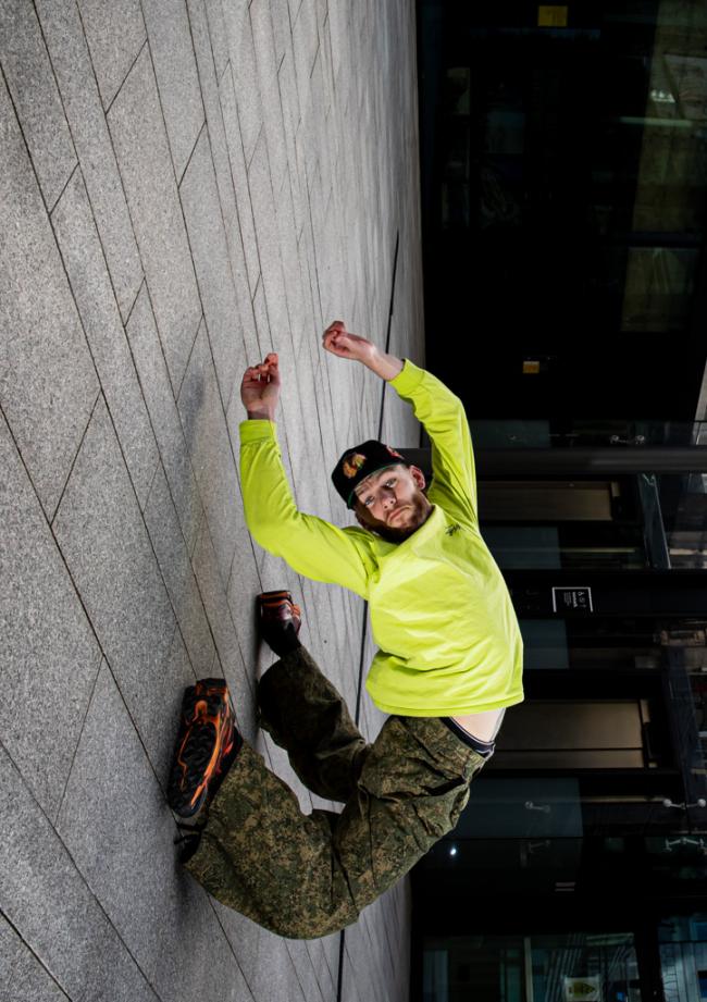 A person in a neon yellow shirt and camo pants mid-jump against a gray stone wall, shot from a tilted angle with city buildings behind.