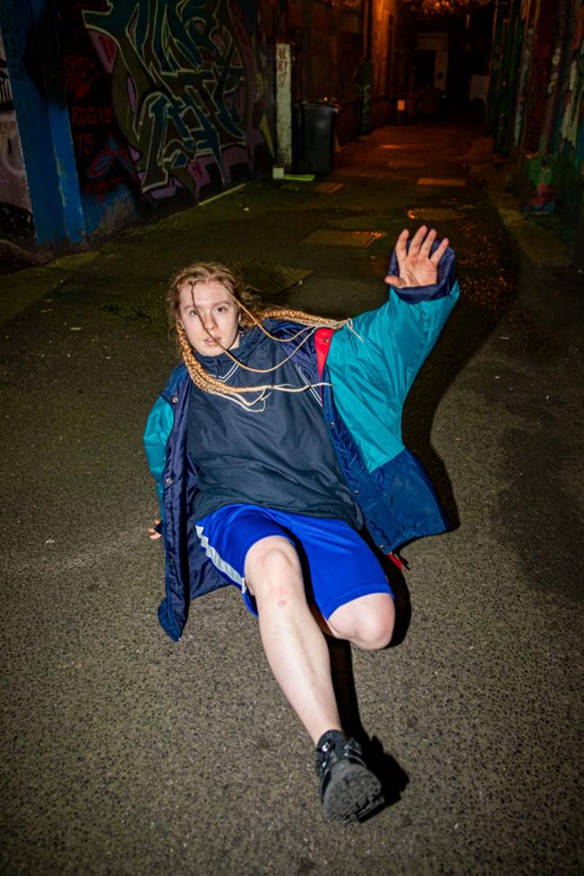 A person with long braids in a blue windbreaker and shorts holds a low floor pose in a graffiti-covered alley at night.
