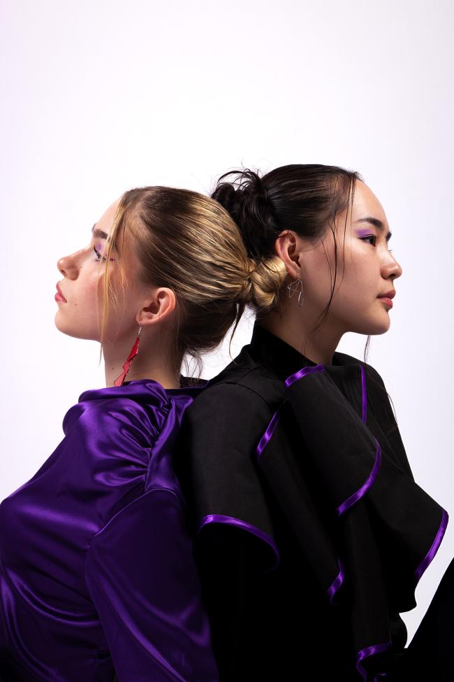 A model wearing a purple silk satin blouse and a second model wearing a black blouse with purple trim