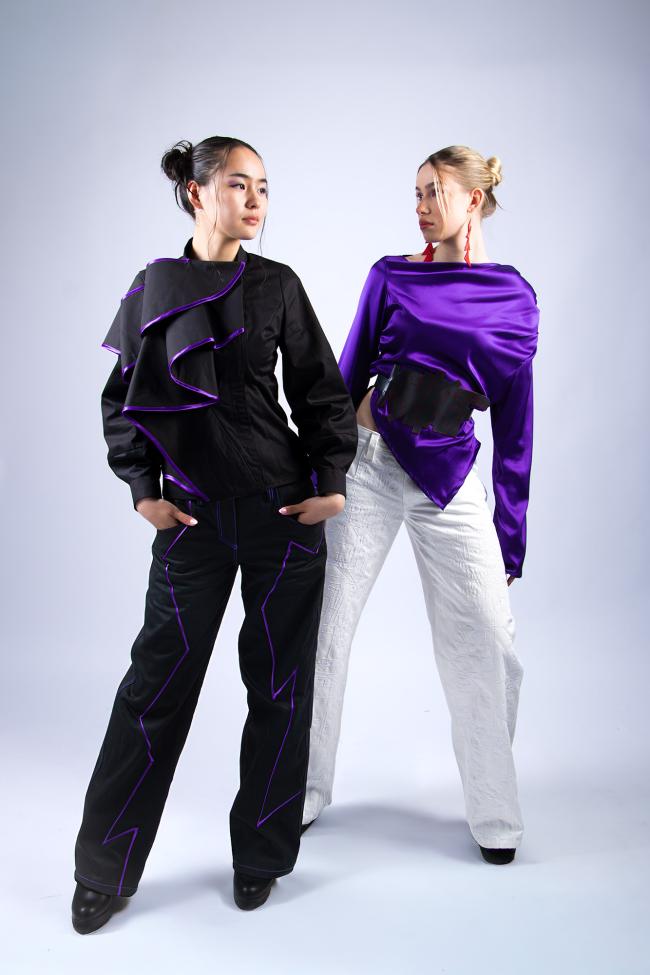 A model wearing a black blouse with purple trim and black trousers, a second model wearing a purple silk satin blouse with white trousers.