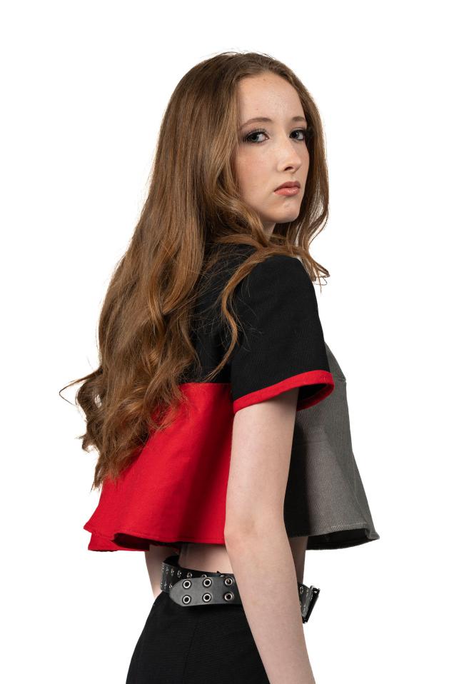 A side view of a female model with long wavy red hair wearing a black, red, and grey top with black trousers.
