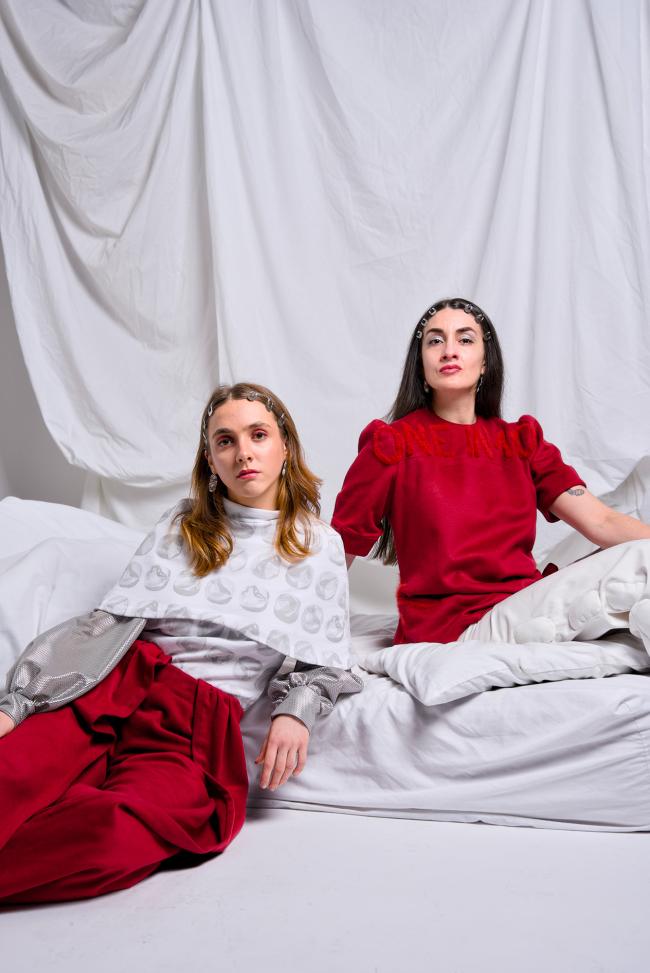 Two models sitting on a bed, contrasting outfits in red and white colours