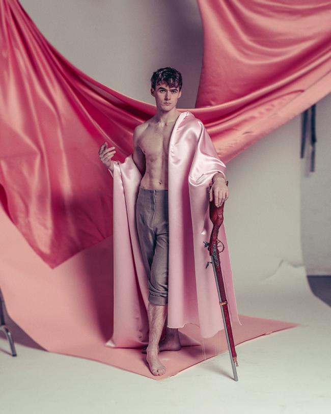  A shirtless figure draped in pink satin stands against flowing fabric, holding an ornate sword in a theatrical studio portrait.