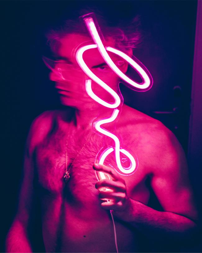 A shirtless figure holds a glowing pink neon light while motion blur obscures parts of the face and body in a dark setting.