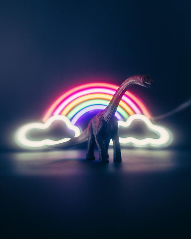 A toy dinosaur stands silhouetted in front of a glowing rainbow neon light in a darkened scene.