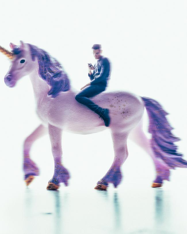 A person sits looking at a phone while riding a large toy unicorn in a surreal, softly lit composite image.