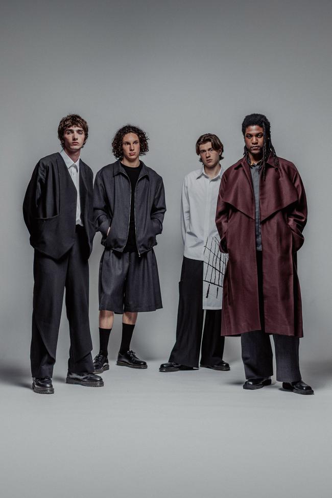 Four young men stand in a minimalist studio against a plain grey background, wearing contemporary, oversized fashion. Their outfits include loose suits, layered shirts, shorts with long socks, and a long maroon coat, all styled in muted tones. They face the camera with neutral expressions, spaced evenly in a slightly staggered formation.