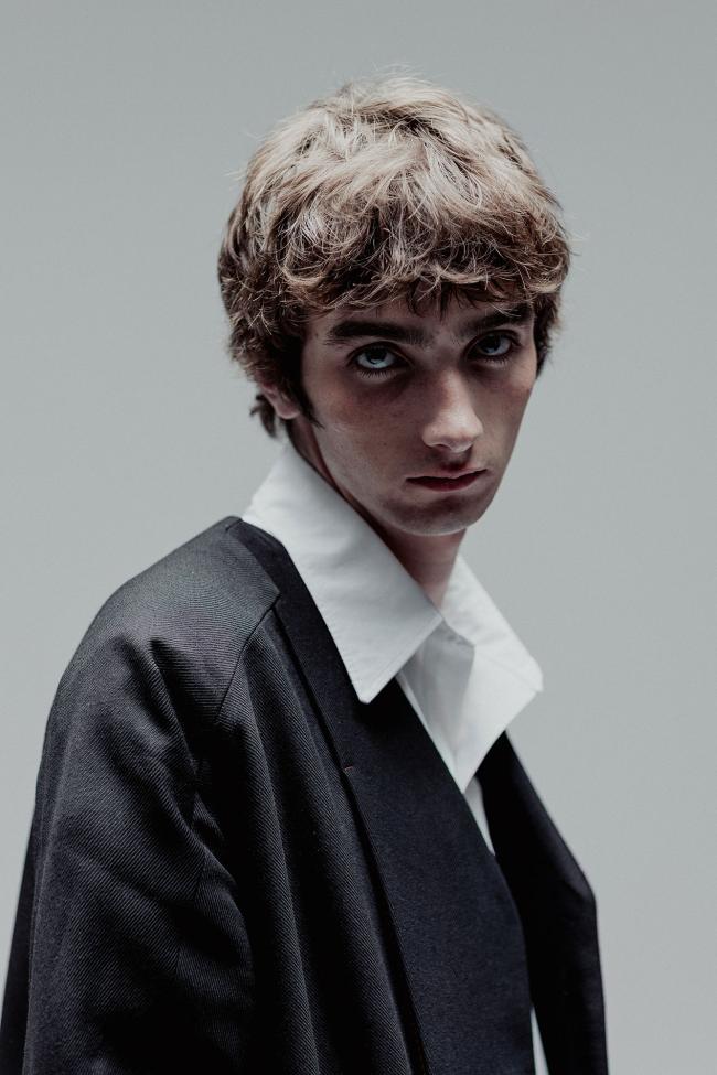 Close-up portrait of a young man with tousled light brown hair, wearing an oversized dark blazer over a white collared shirt. He faces slightly to the side while looking directly at the camera with a neutral, serious expression, set against a plain grey background.