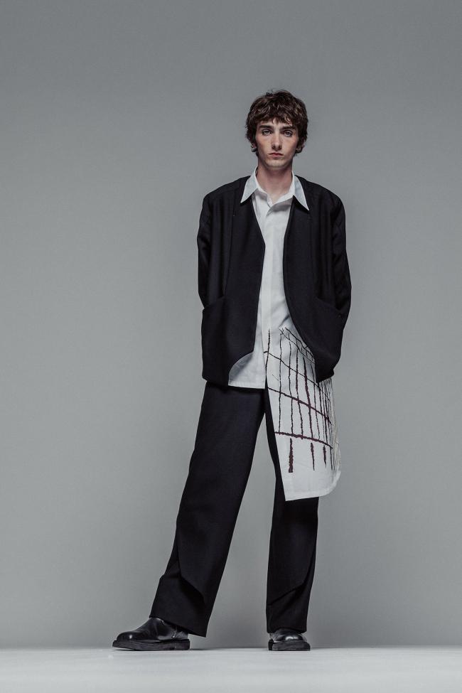 Full-body studio portrait of a young man standing against a plain gray background, wearing an oversized black suit with wide-leg trousers and a white shirt featuring an abstract linear pattern. He faces forward with a neutral expression, hands behind his back, and one leg slightly bent.