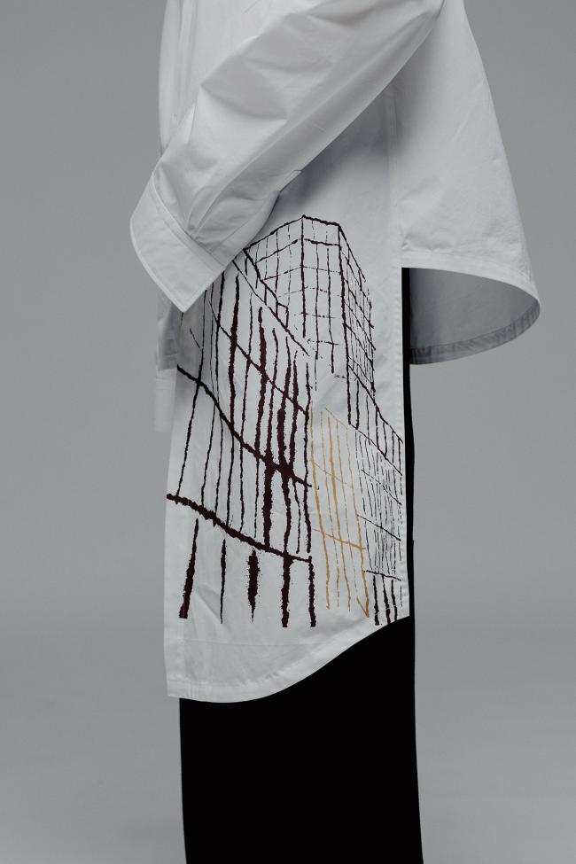  A detailed close-up shot of the lower side seam and hem of a white button-up shirt worn over black pants, against a plain grey background. The shirt features an architectural line-art print on the side panel, rendered in dark burgundy and muted gold. A diagonal panel of the white shirt extends downwards below the main hem.