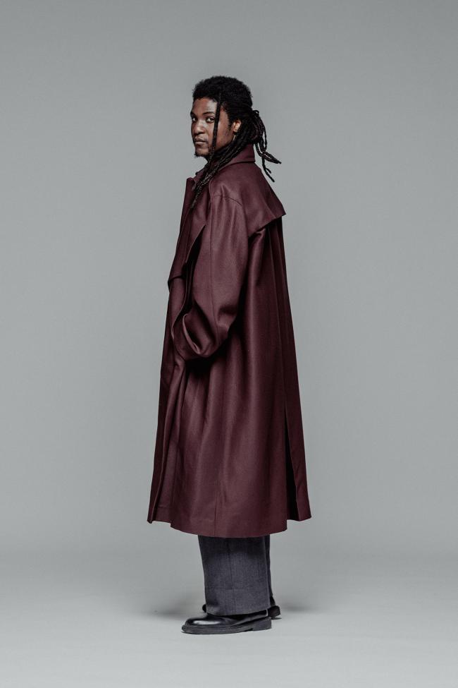  A full-length fashion portrait of a man with dark dreadlocks looking over his shoulder against a solid grey background. He is wearing a voluminous, oversized burgundy trench coat with a storm flap on the back, paired with wide-leg charcoal grey trousers and chunky black leather boots.