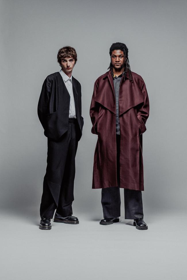 A full-length fashion portrait of two men standing side-by-side against a solid grey background. The man on the left wears a structured, collarless black blazer over a white button-up shirt, paired with wide-leg black trousers and chunky black boots. The man on the right wears an oversized, voluminous burgundy trench coat over a grey shirt, charcoal wide-leg trousers, and black leather boots. 