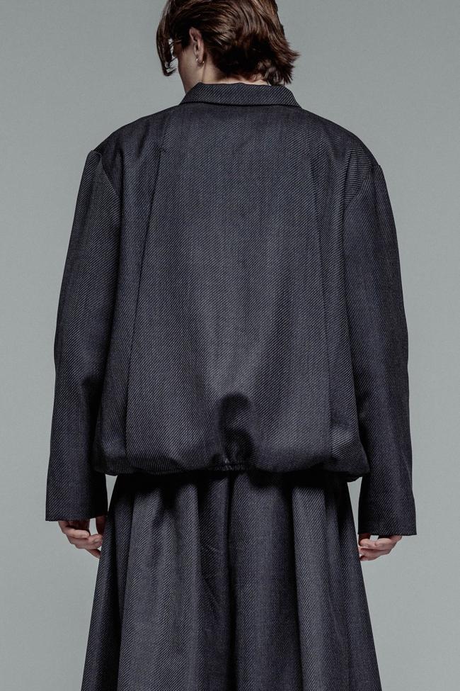 A medium shot from the back of a person with shoulder-length brown hair, wearing a structured, charcoal grey textured jacket and matching voluminous pleated trousers or skirt. The jacket has a gathered, elasticated hem that creates a rounded silhouette, set against a plain grey background.