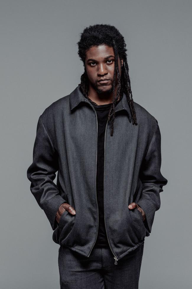  A medium fashion portrait of a man with dark dreadlocks standing against a solid grey background. He is wearing a structured, dark grey textured zip-up jacket with a collar and his hands in his pockets. The jacket is worn over a black shirt and paired with matching grey trousers.