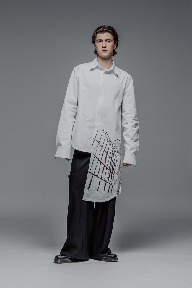 A full-length fashion portrait of a man with brown hair standing against a solid grey background. He is wearing a white button-up shirt with an asymmetrical hem; a long rectangular panel on the left side features a burgundy and gold architectural line-art print. The look is paired with oversized, wide-leg black trousers and chunky black leather boots. 