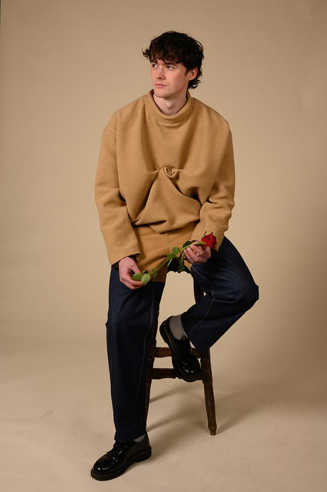 Model sits on stool wearing an oversized camel crewneck and a pair of jeans while holding a rose in front of a beige background