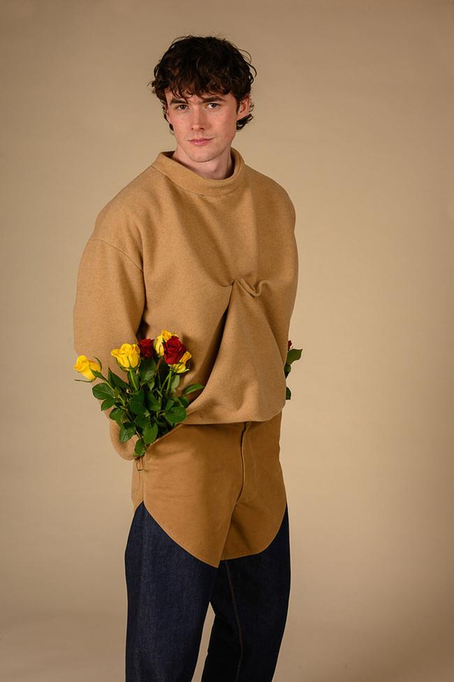 Model stands in studio wearing camel crewneck with denim a suede jeans with red and yellow roses placed inside jeans pockets
