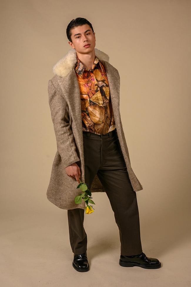 Male model stands in studio wearing a long grey herringbone overcoat with a floral shirt, holding a yellow rose in front of a beige background