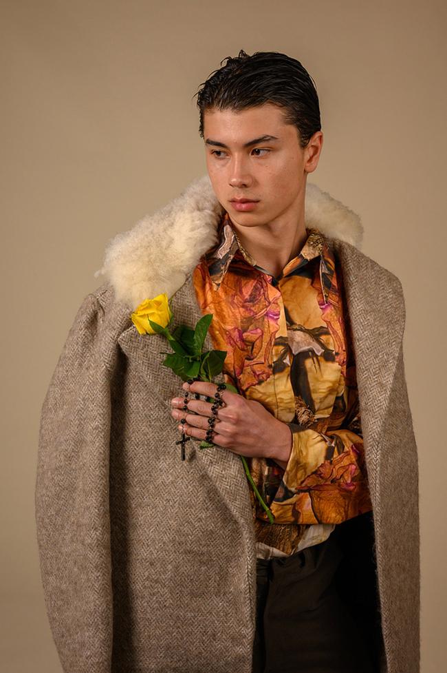 Male model in herringbone overcoat with sheepskin fur collar and floral shirt, holds a yellow rose against a beige background