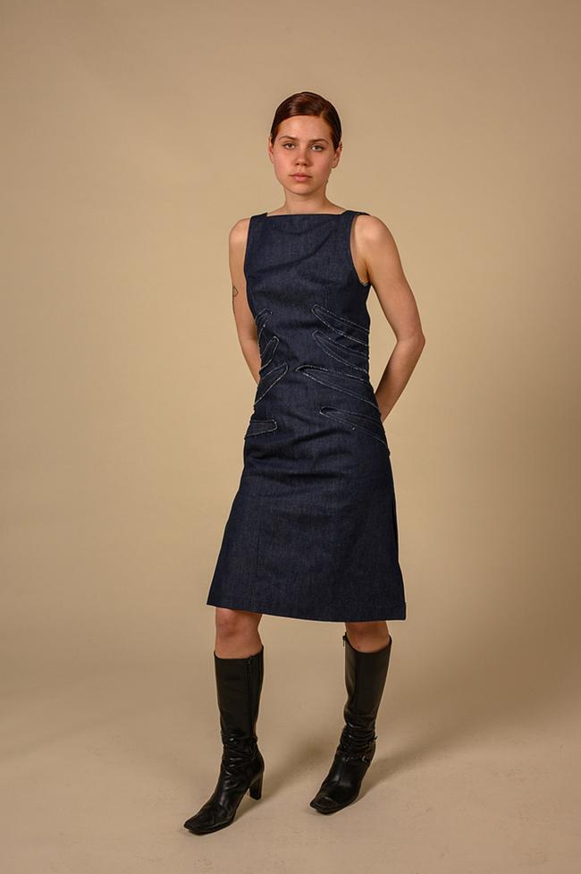 A female model stands in a studio wearing a dark blue denim dress and black boots against a beige background.