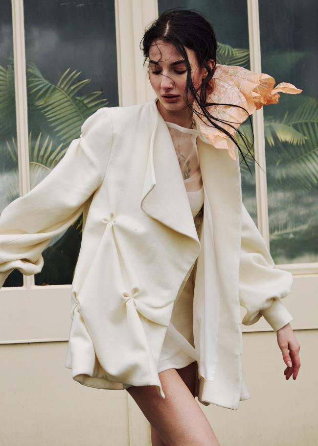Close-up of a model in a short, cream wool coat standing against a greenhouse background.