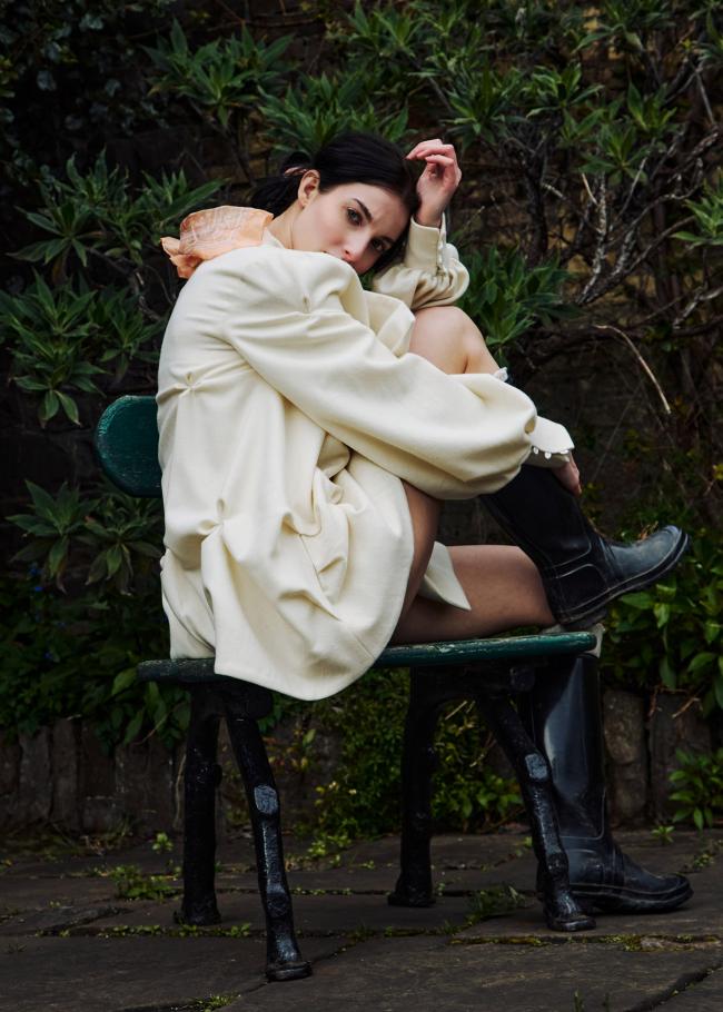  Model in a short cream coat with flower fabric manipulation, sitting on a bench with one leg up.