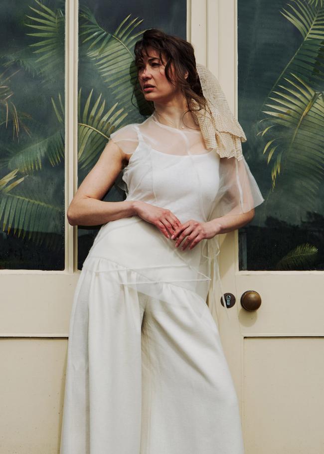 Model in sheer top and gathered trousers against a greenhouse background.