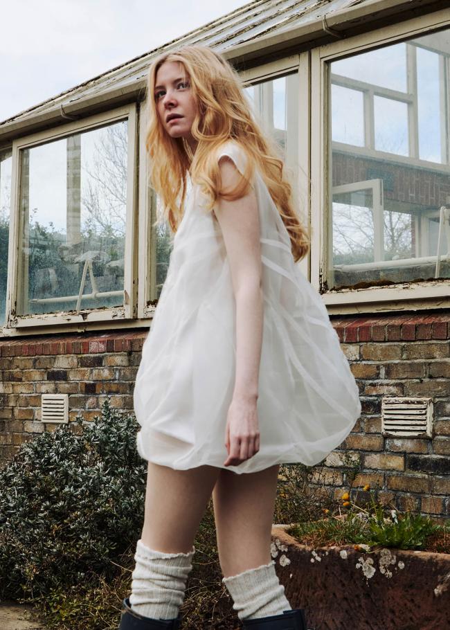 Model in bubble dress walking away and looking to the side with a small greenhouse in the background.