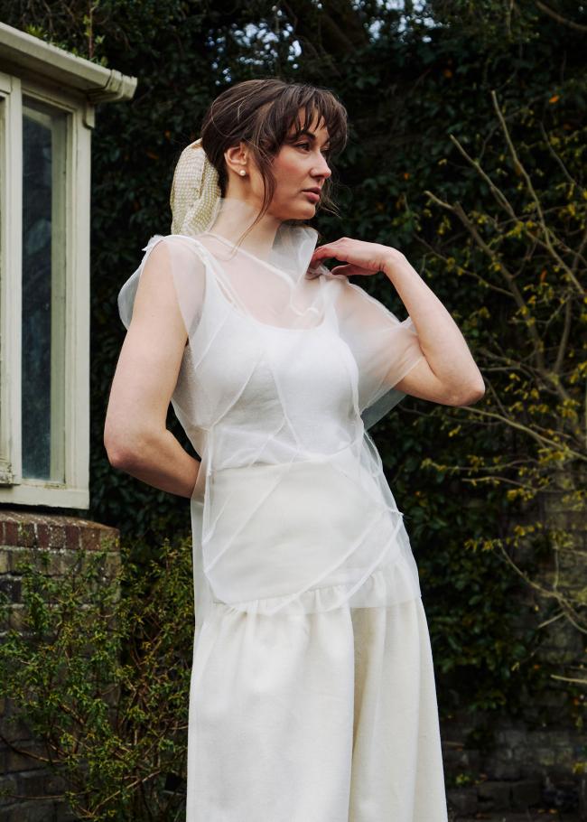 Model in a sheer top with raw seamlines and gathered trousers standing with her hand on her shoulder. 