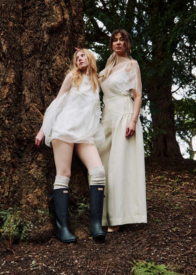 Two models standing close together against a tree. One in a sheer bubbly dress and the other in a sheer top and gathered wide-leg trousers.